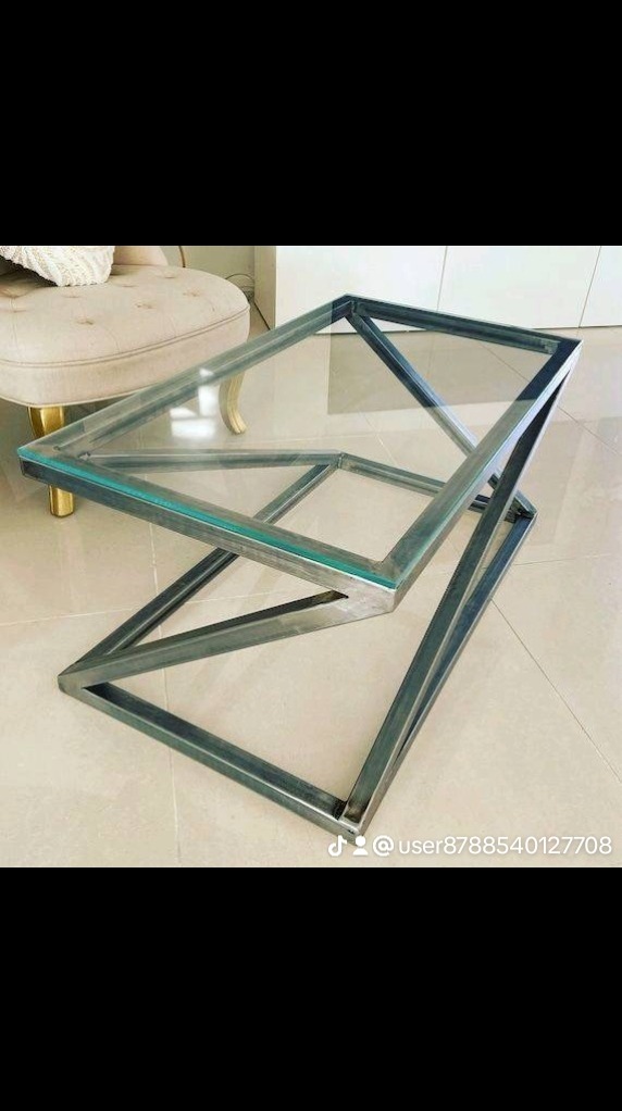 Szklany stolik kawowy z geometryczną, metalową podstawą, ustawiony na beżowej podłodze, z widocznym fragmentem kremowego fotela w tle.