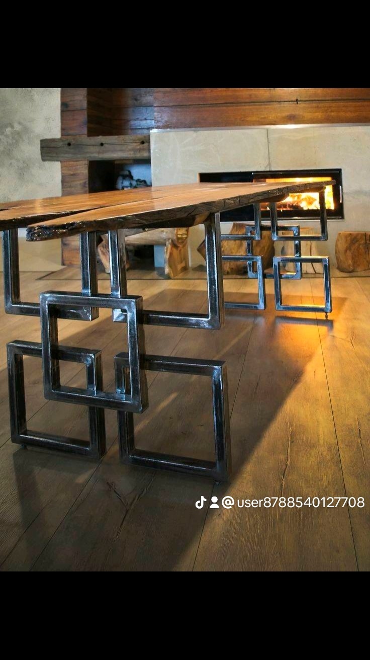 Stół z blatem z surowego drewna i metalową podstawą z kwadratowych elementów, widok z dołu, w tle kominek z palącym się ogniem.