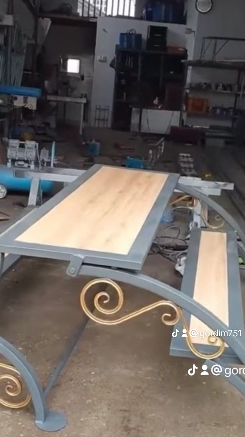 Stół z ławką o metalowej konstrukcji w odcieniach szarości z dekoracyjnymi złotymi elementami, blat i siedzisko z jasnego drewna, w tle warsztat z różnymi narzędziami i maszynami.