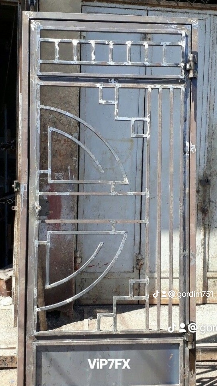 Surowa, metalowa brama z geometrycznymi zdobieniami w trakcie spawania, widoczne ślady obróbki metalu i niedoskonałości powierzchni.