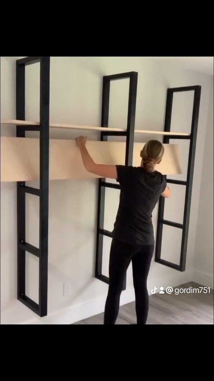 Kobieta montuje jasną półkę w czarnym, metalowym regale drabinkowym na białej ścianie w pomieszczeniu.