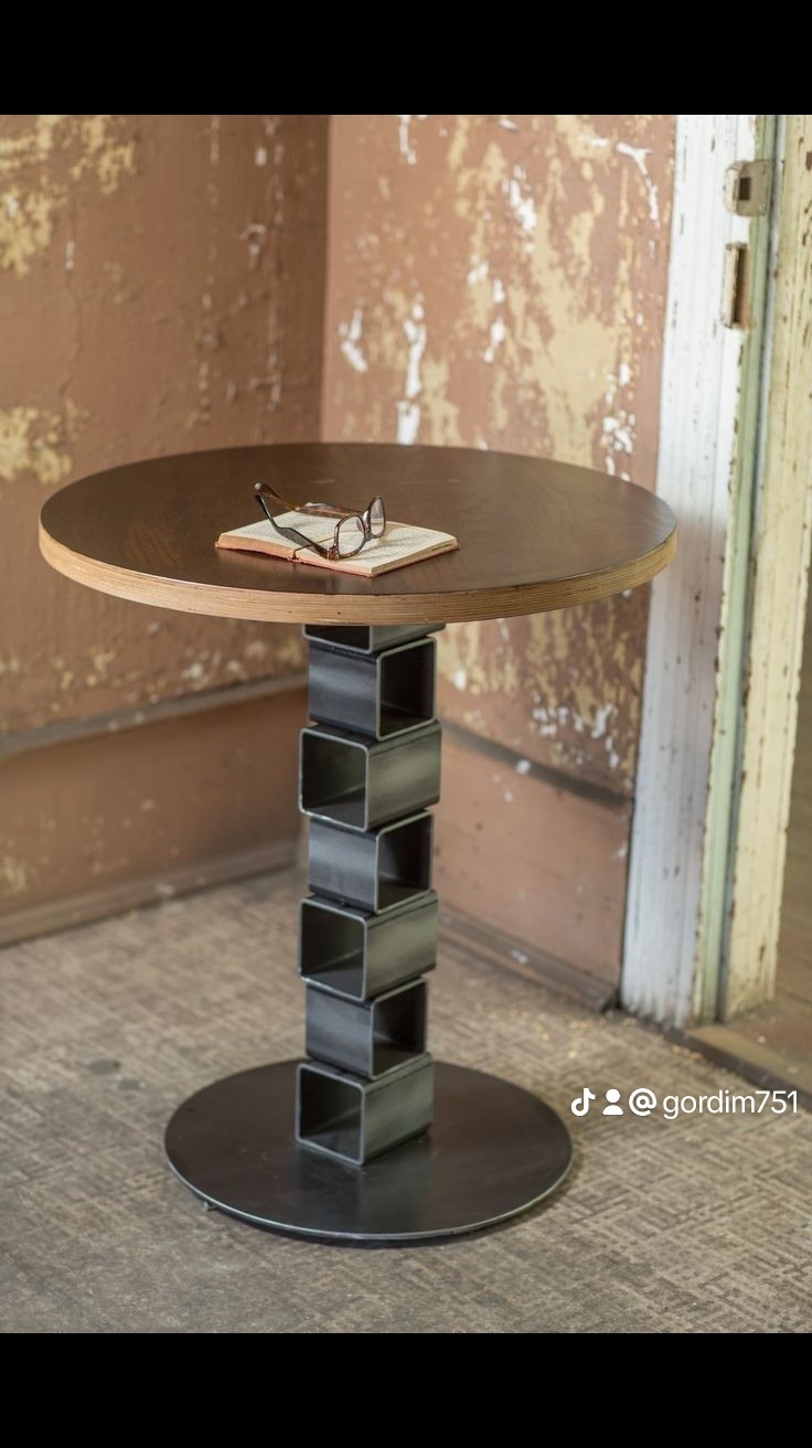 Okrągły stolik kawowy z blatem z ciemnego drewna i metalową podstawą z kwadratowych elementów, na blacie leżą okulary i notatnik, tło stanowią ściana z łuszczącą się farbą i fragment drzwi.