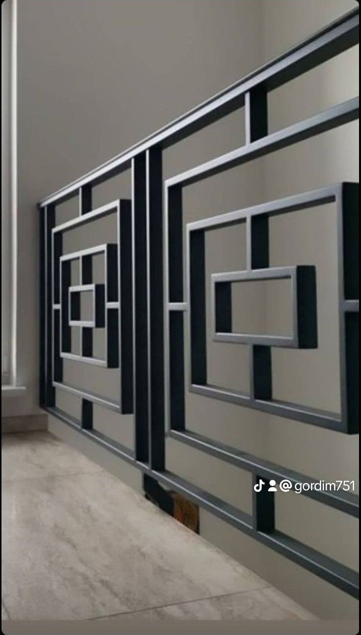 Nowoczesna, metalowa balustrada schodowa z geometrycznym wzorem kwadratów, pomalowana na ciemnoszary kolor, na tle ściany i podłogi w odcieniach szarości.