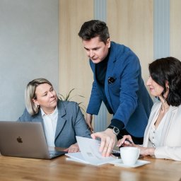 ONE ESTATE Doradca Kredytowy Kraków - Trzy osoby w biurze omawiają dokumenty przy biurku z laptopem i filiżanką kawy. Mężczyzna w niebieskiej marynarce stoi, wskazując na dokument.