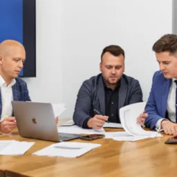 Trzech mężczyzn w garniturach analizuje dokumenty i laptopa przy drewnianym stole w biurze, z logo firmy nieruchomości na ekranie w tle.