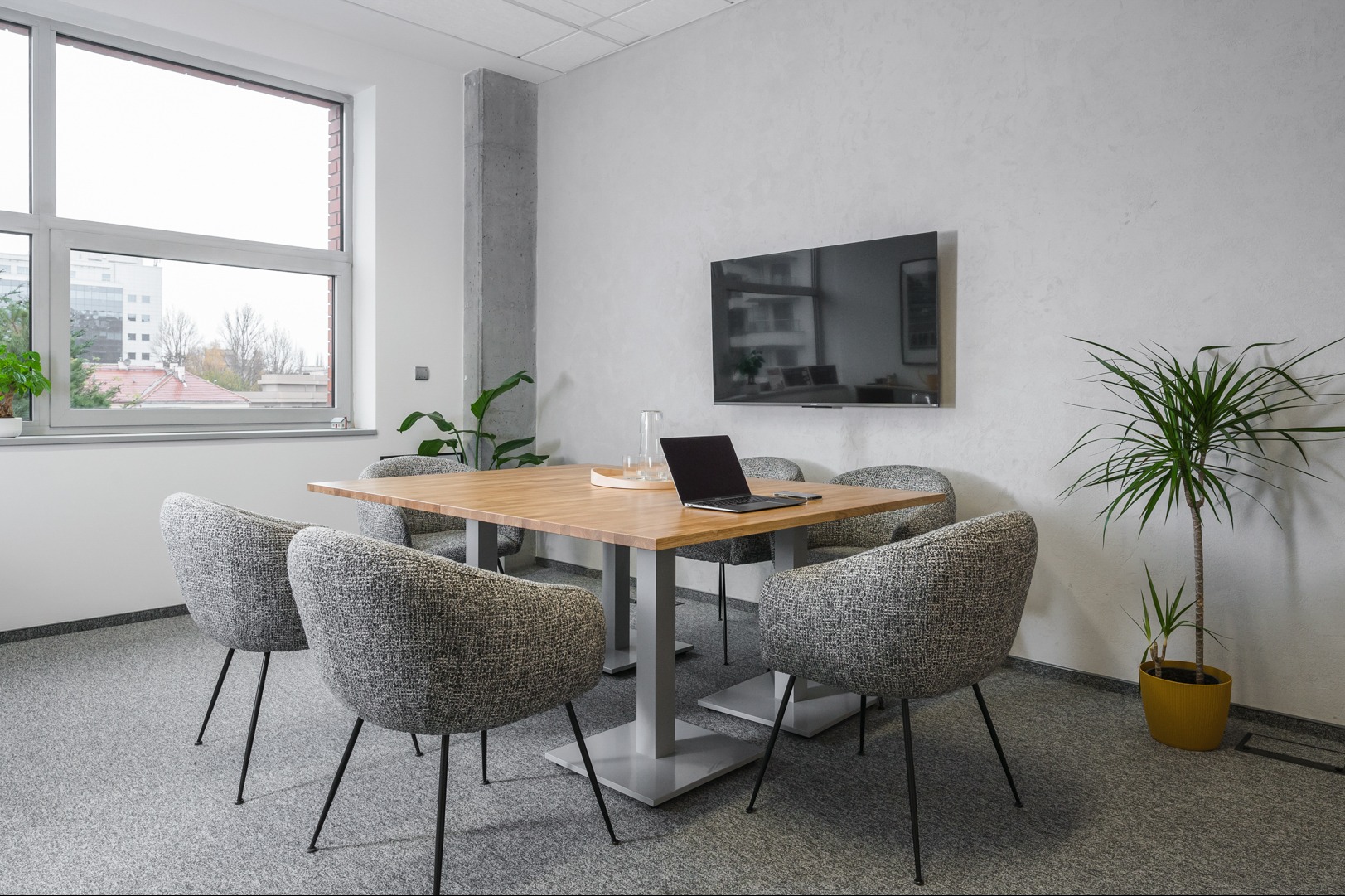 Jasne biuro z drewnianym stołem konferencyjnym, szarymi krzesłami i laptopem. Minimalistyczny design, roślina w doniczce i telewizor na ścianie. Widok z okna na budynki.