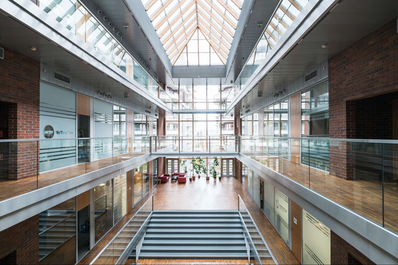 Wnętrze nowoczesnego biurowca z atrium, widok z góry na schody, szklane balustrady i biura. Duże okna wpuszczają naturalne światło. Widoczne logo firmy na szklanych ścianach.