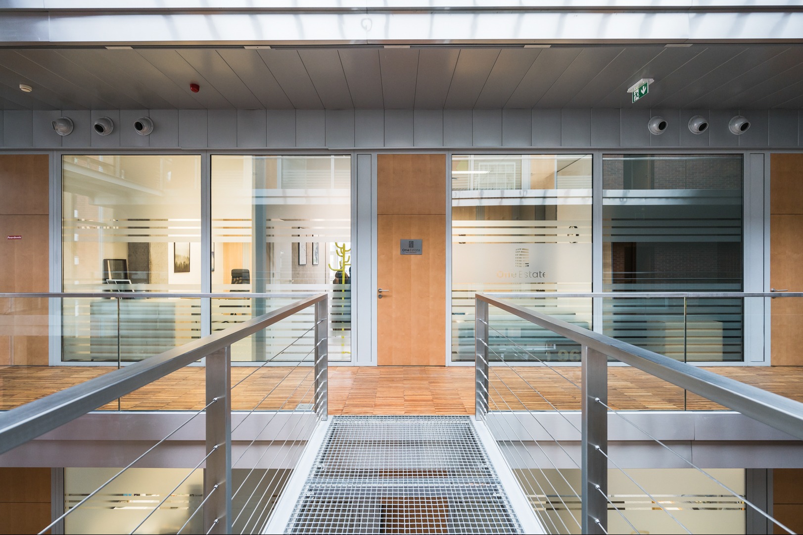 Wnętrze biura z metalową kładką, szklanymi ścianami i drewnianą podłogą. Widoczne logo 'OneEstate' na drzwiach i szybach. Jasne oświetlenie.