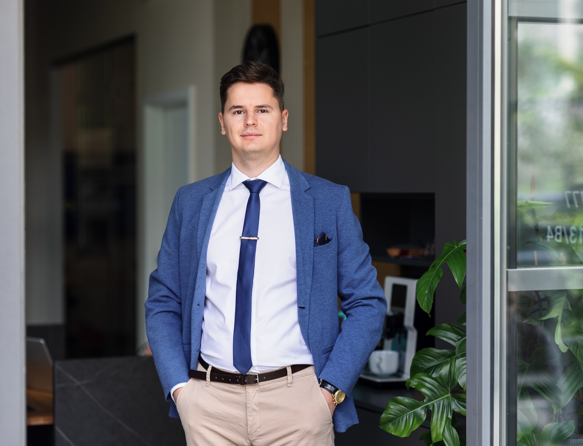 Mężczyzna w błękitnej marynarce i krawacie, stojący w nowoczesnym wnętrzu z roślinami, prezentujący profesjonalny wizerunek.