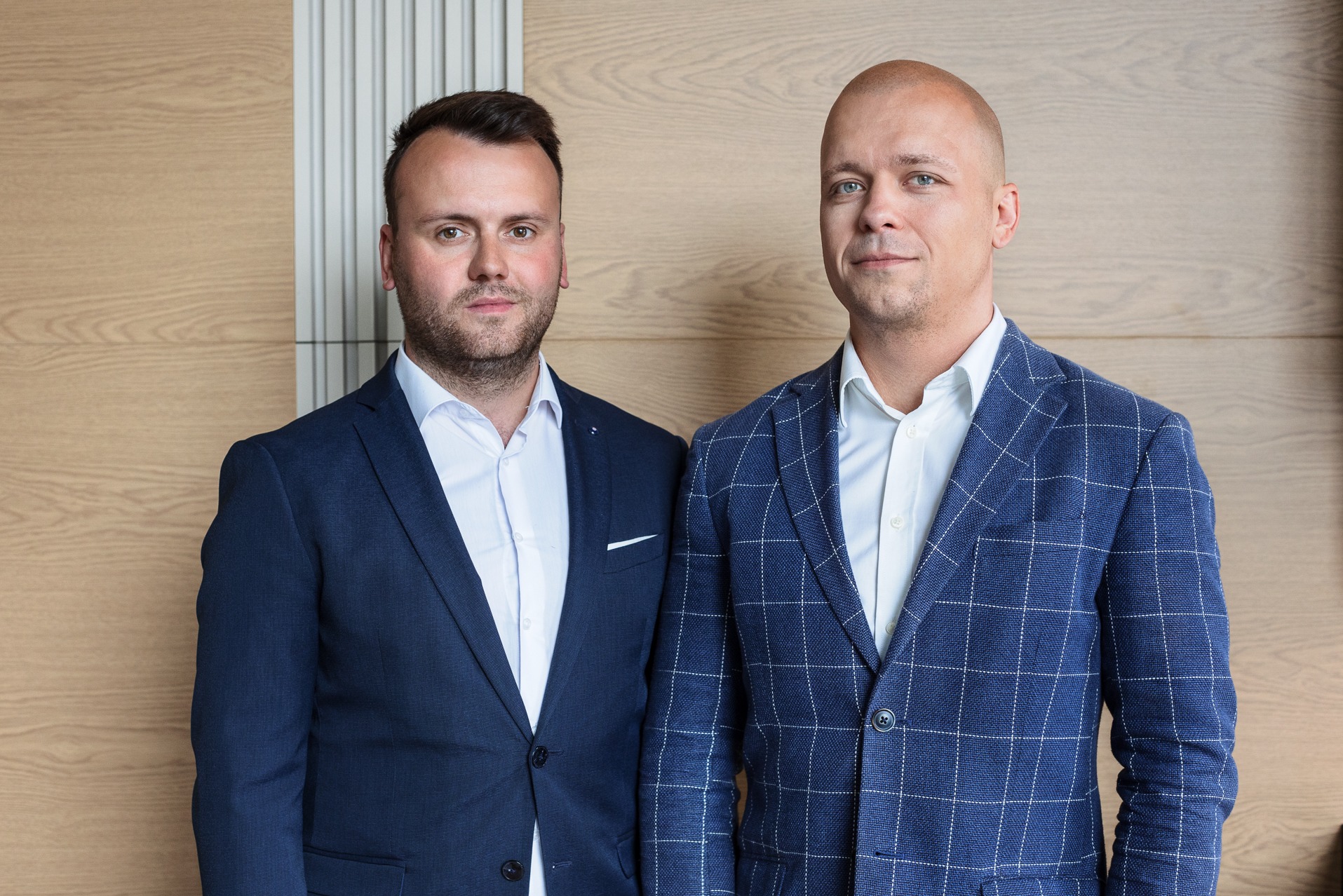 Portret dwóch mężczyzn w garniturach na tle drewnianej ściany. Jeden w gładkim, granatowym garniturze, drugi w niebieskim garniturze w kratę. Profesjonalny wygląd.