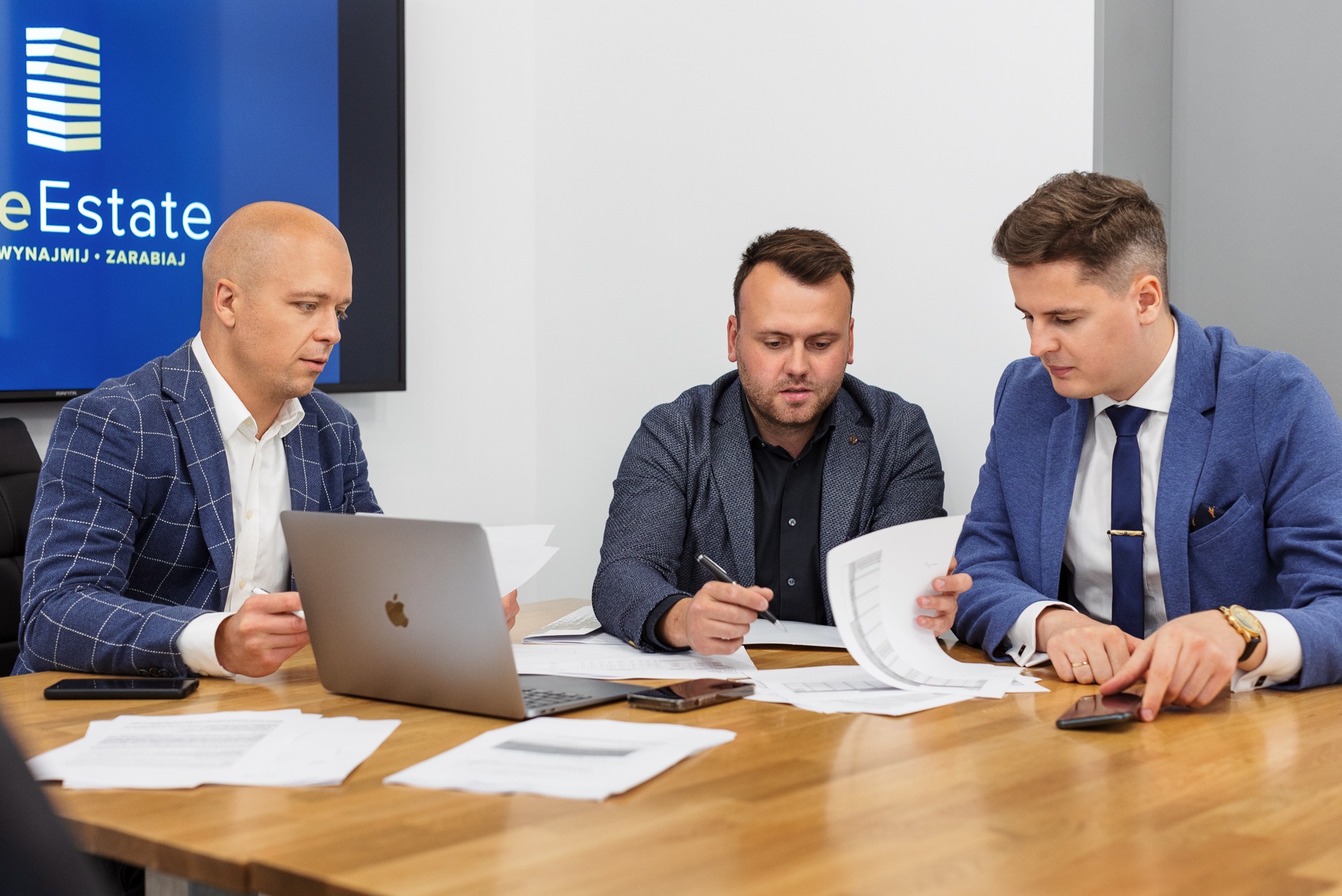 Spotkanie biznesowe: trzech mężczyzn w garniturach analizuje dokumenty przy drewnianym stole z laptopem i smartfonami, logo firmy na ekranie w tle.