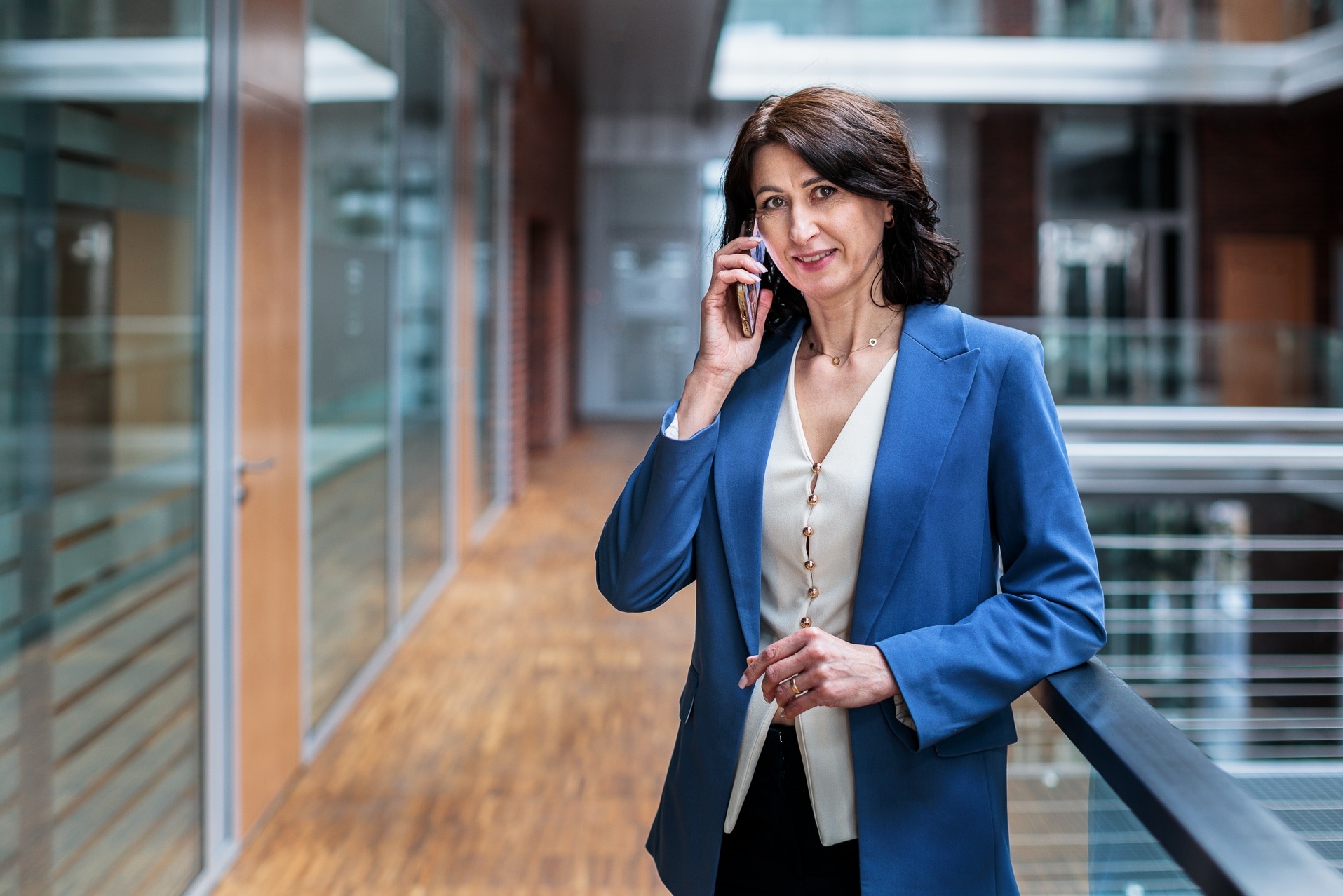 Kobieta w niebieskiej marynarce rozmawia przez telefon w nowoczesnym biurze. Widoczne szklane ściany i metalowa balustrada. Uśmiech na twarzy, profesjonalny wygląd.