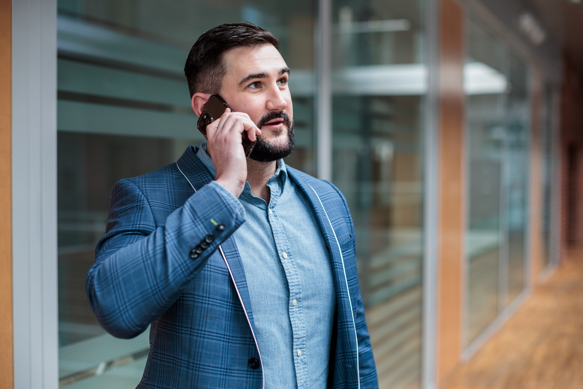 Mężczyzna w niebieskiej marynarce w kratę rozmawia przez telefon w nowoczesnym biurze z przeszklonymi ścianami. Uśmiechnięty, patrzy w górę.
