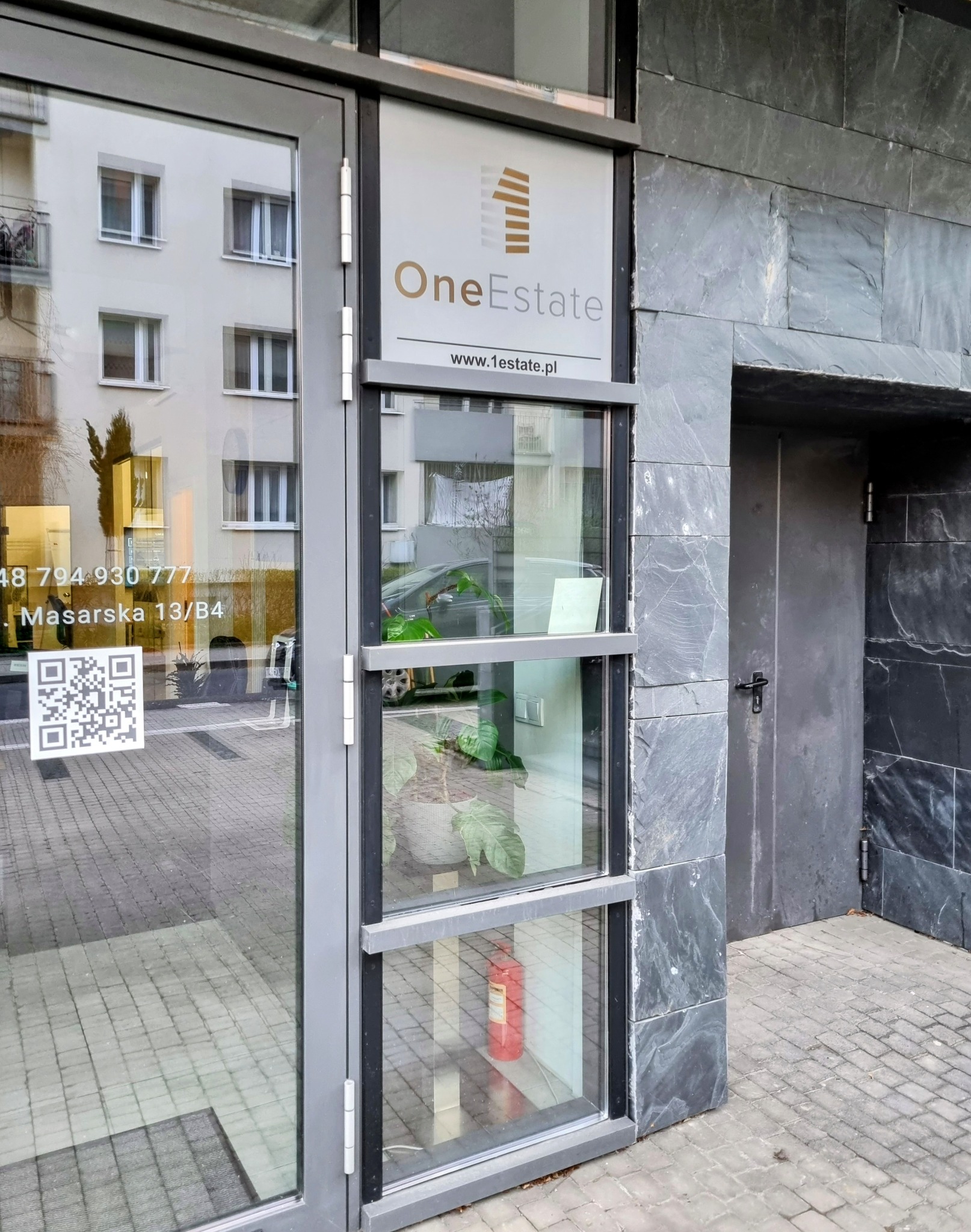 Wejście do biura agencji nieruchomości One Estate z logo, adresem Masarska 13/B4 i kodem QR na szybie, odbicie budynku w szkle.