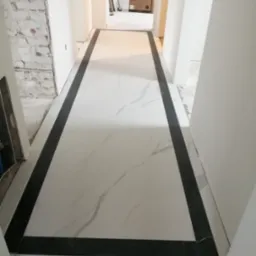 Nowo ułożona podłoga z białych płytek imitujących marmur, obramowana czarnymi pasami, widoczna w korytarzu w trakcie remontu, z fragmentem surowej ściany z cegły po lewej stronie.