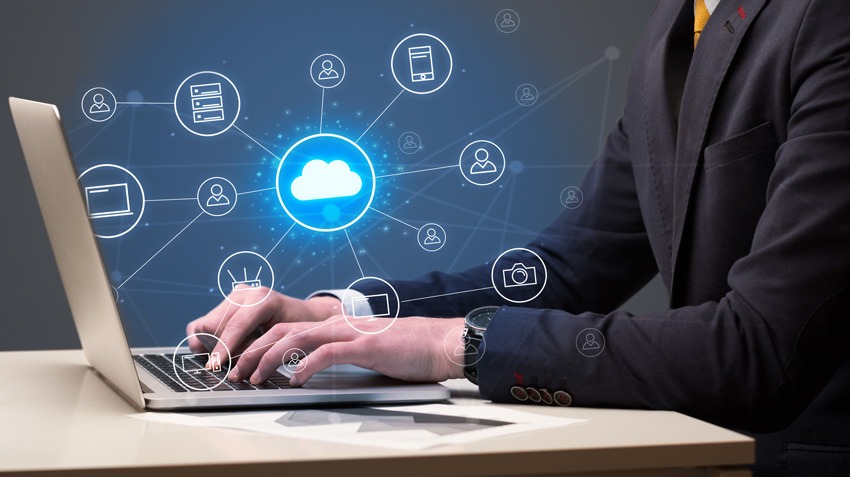 Mężczyzna w garniturze pisze na laptopie z nałożoną grafiką chmury i ikon urządzeń, symbolizującą połączenie z siecią i dostęp do danych.