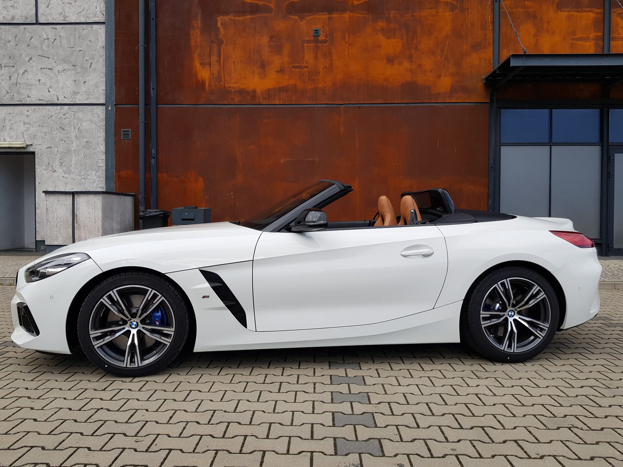Biały kabriolet BMW Z4 z brązowymi skórzanymi siedzeniami, zaparkowany na brukowanej nawierzchni przed budynkiem z rustykalną, rdzawą ścianą i szarą, betonową ścianą.