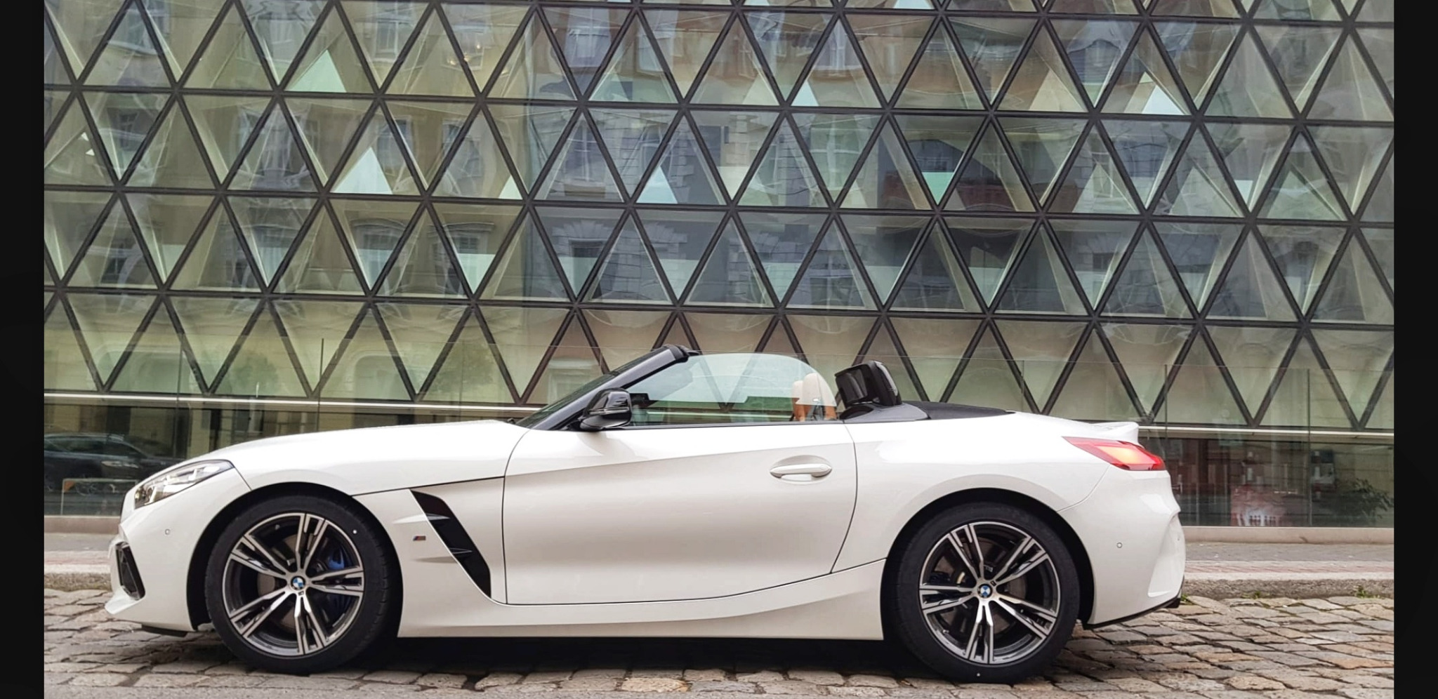 Biały kabriolet zaparkowany na brukowanej ulicy z nowoczesnym budynkiem w tle, którego fasada składa się z trójkątnych okien.