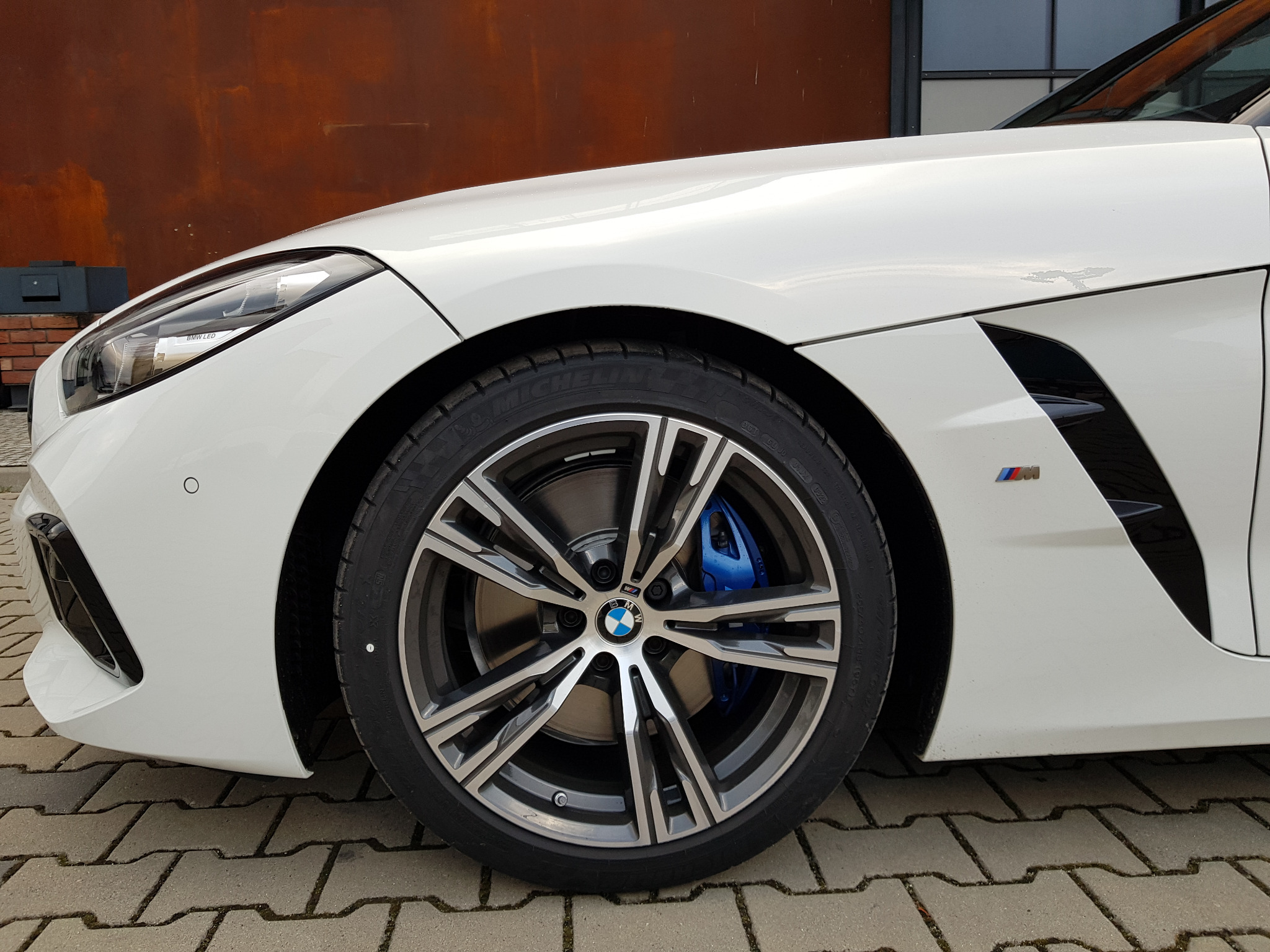 Fragment białego samochodu BMW Z4 z widocznym przednim kołem, niebieskimi zaciskami hamulcowymi M Performance i emblematem M na błotniku, na tle kostki brukowej i metalowej ściany.