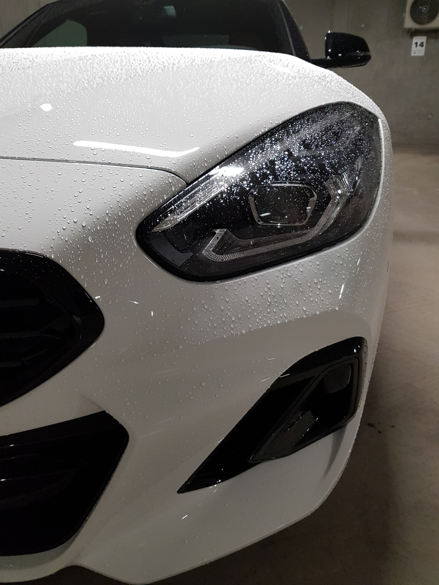Biały samochód z kroplami wody na karoserii i reflektorze, widok z przodu na fragment maski i lampę w garażu podziemnym.