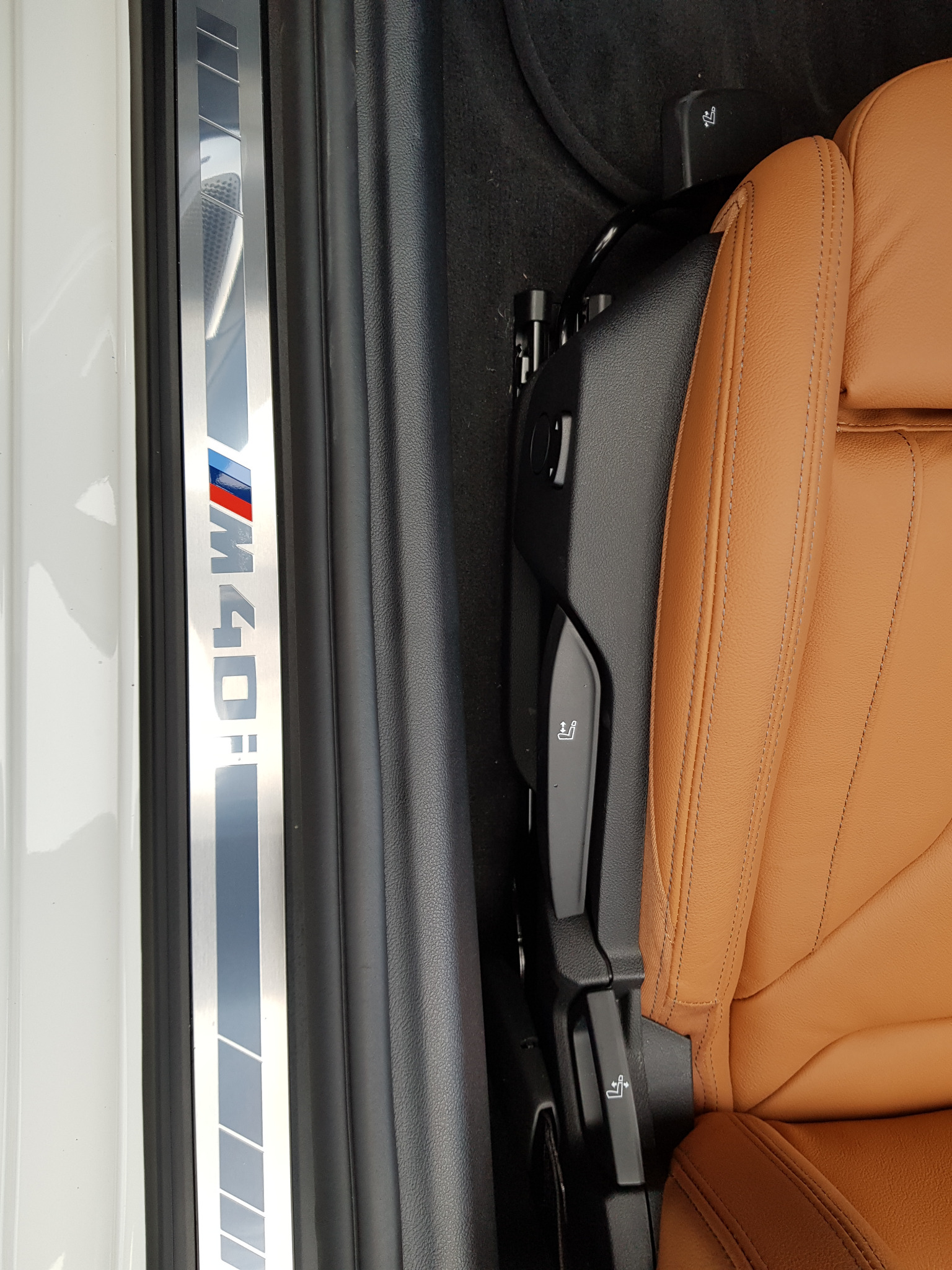 Wnętrze samochodu z brązową skórzaną tapicerką fotela i podświetlaną listwą progową z napisem M40i, widoczne elementy regulacji fotela.