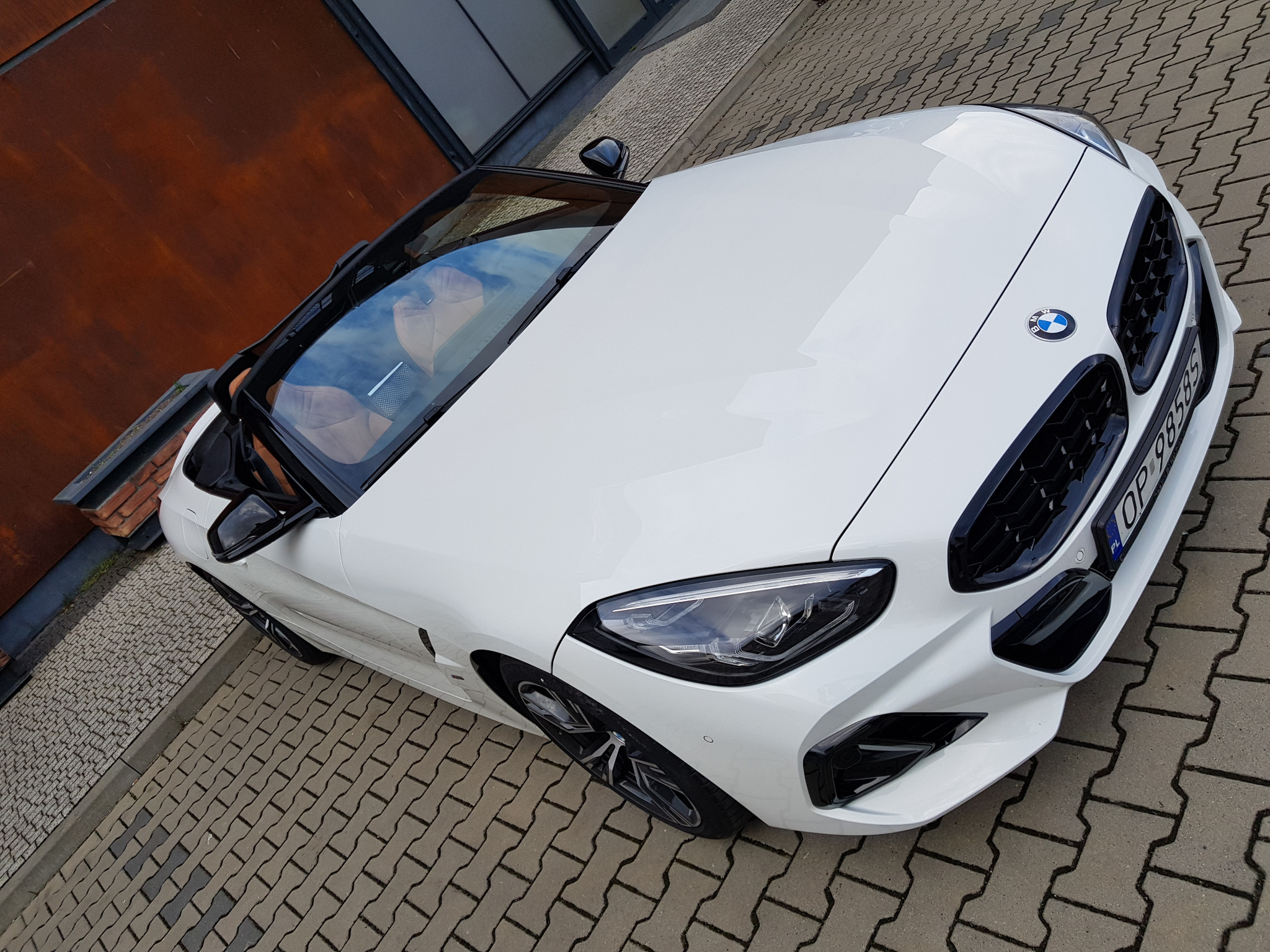 Biały kabriolet BMW Z4 z opolską rejestracją zaparkowany na kostce brukowej przed nowoczesnym budynkiem.