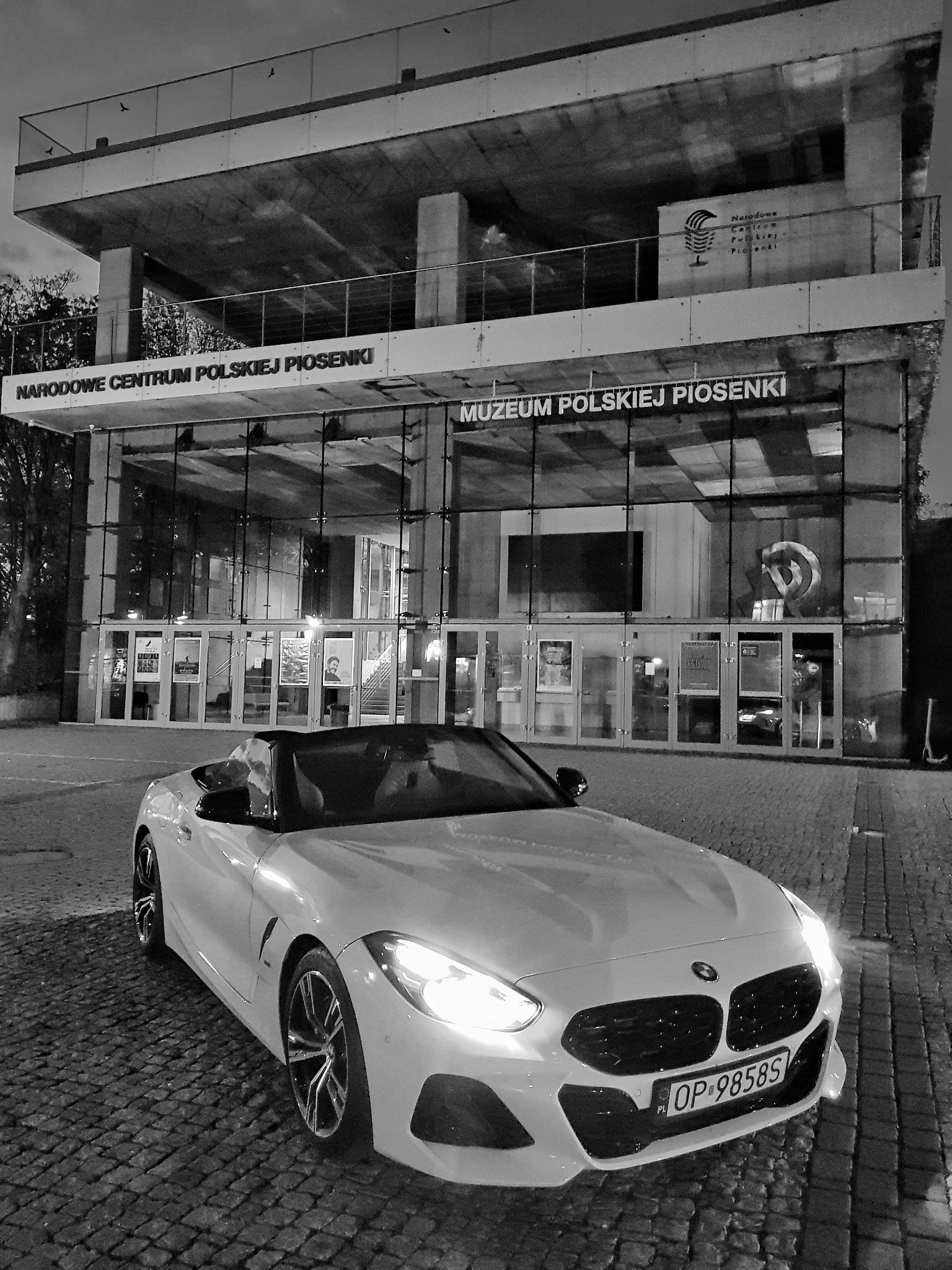 Biały kabriolet BMW Z4 zaparkowany nocą przed Muzeum Polskiej Piosenki w Opolu; widoczne oświetlone wejście do muzeum i bruk uliczny.