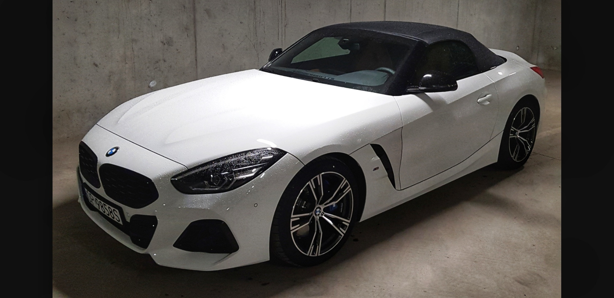 Biały kabriolet BMW Z4 z czarnym dachem, pokryty kroplami wody, zaparkowany w garażu podziemnym.