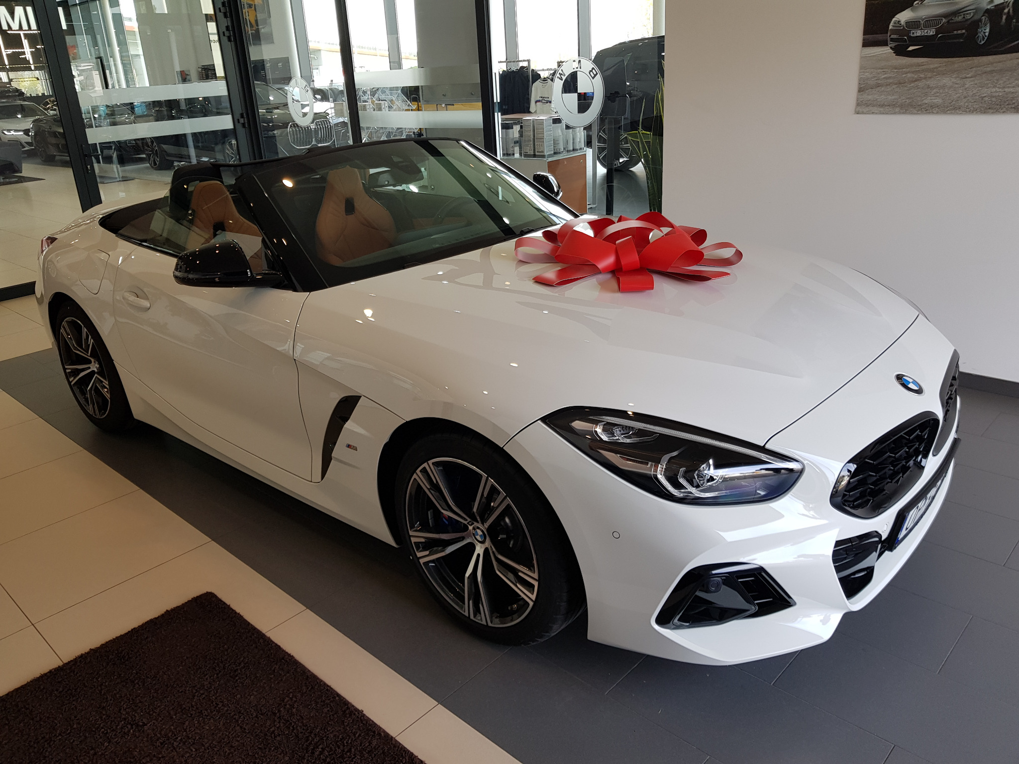 Biały kabriolet BMW Z4 z czerwoną kokardą na masce prezentowany w salonie samochodowym, widoczne beżowe skórzane wnętrze i felgi aluminiowe.