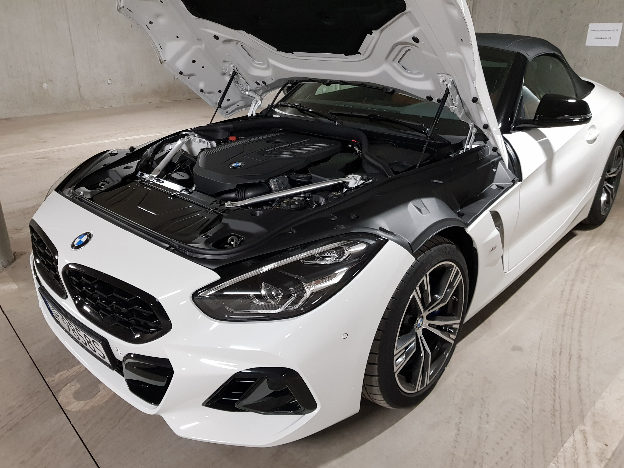 Biały sportowy samochód z otwartą maską, widoczny silnik, zaparkowany w garażu podziemnym.