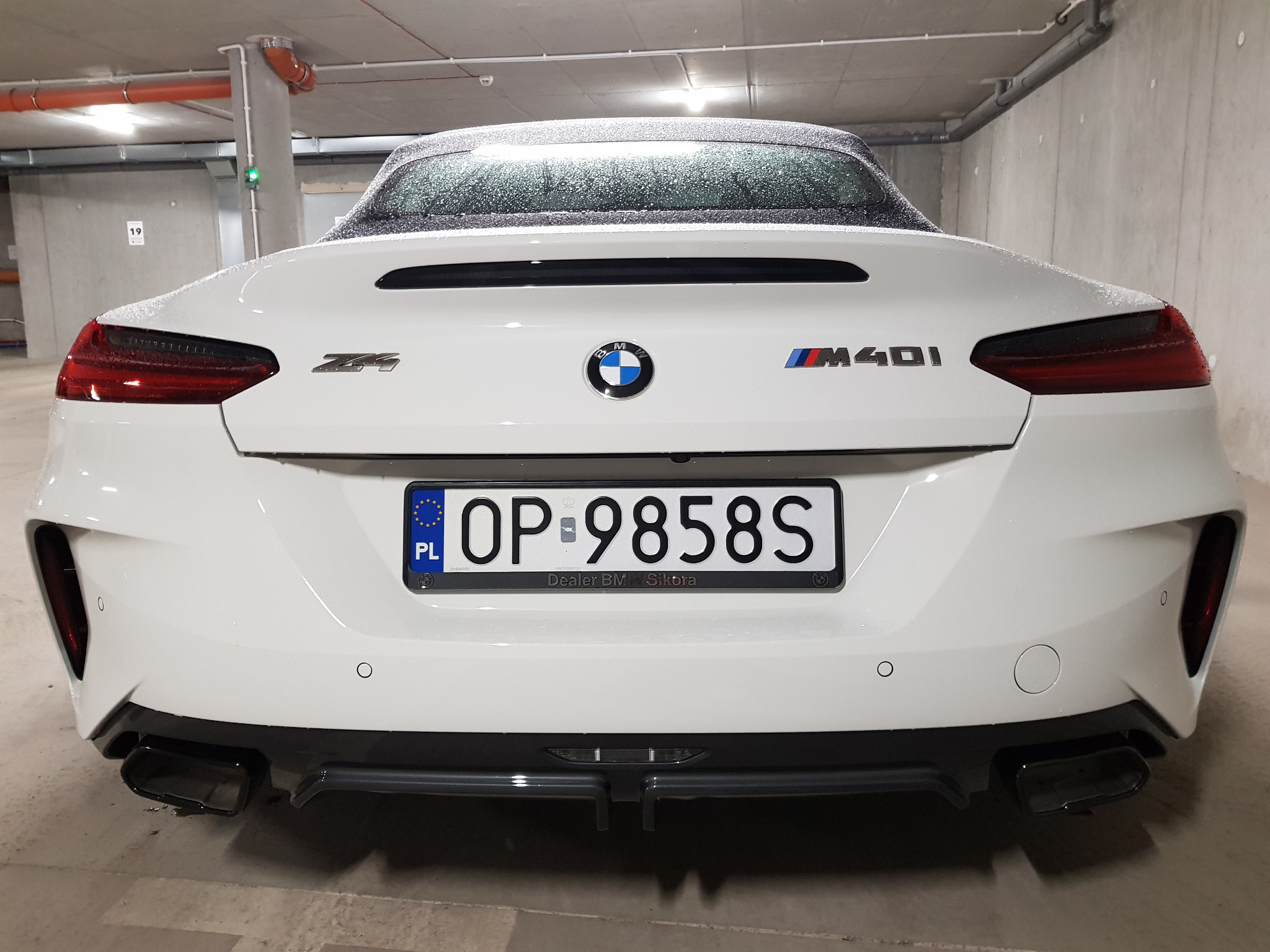 Tył białego BMW Z4 M40i, widoczne tablice rejestracyjne OP 9858S z naklejką dealera BMW Sikora, zaparkowany w garażu podziemnym, krople wody na karoserii.