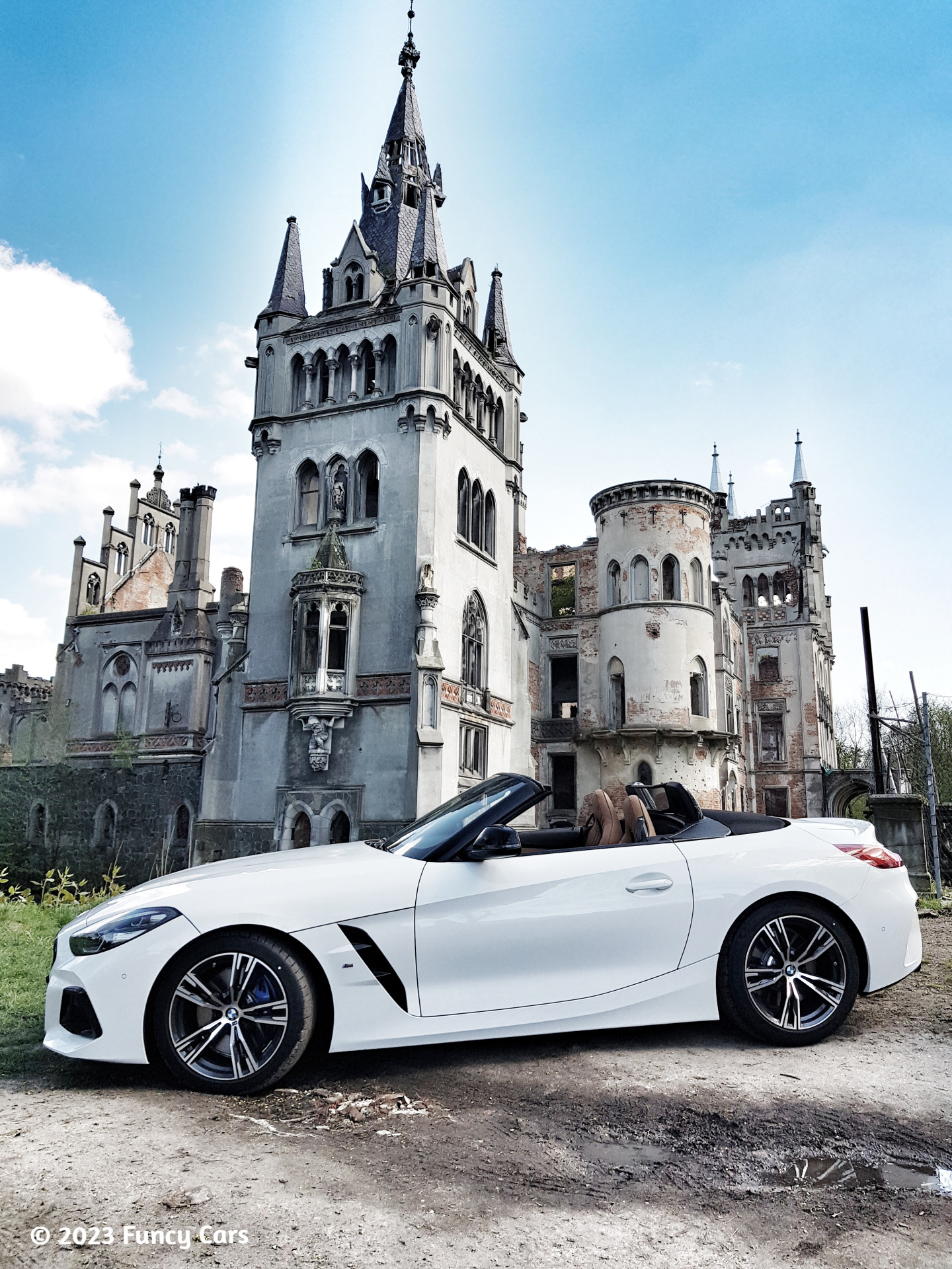Biały kabriolet BMW Z4 zaparkowany na tle malowniczego, choć zrujnowanego gotyckiego zamku z wieżami i detalami architektonicznymi.