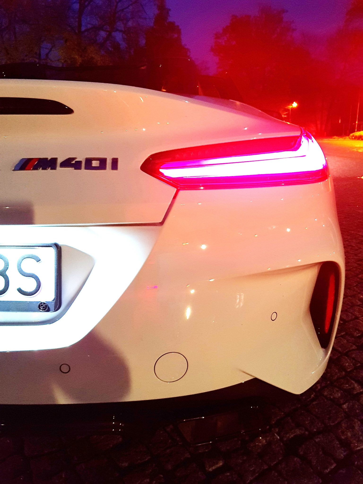 Biały samochód marki BMW M40i, widok z tyłu, oświetlony czerwonym światłem ulicznym, nocna sceneria.