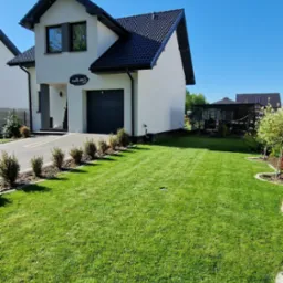 Zadbany trawnik przed nowoczesnym domem z ciemną stolarką okienną i garażem, w tle widoczna altana.