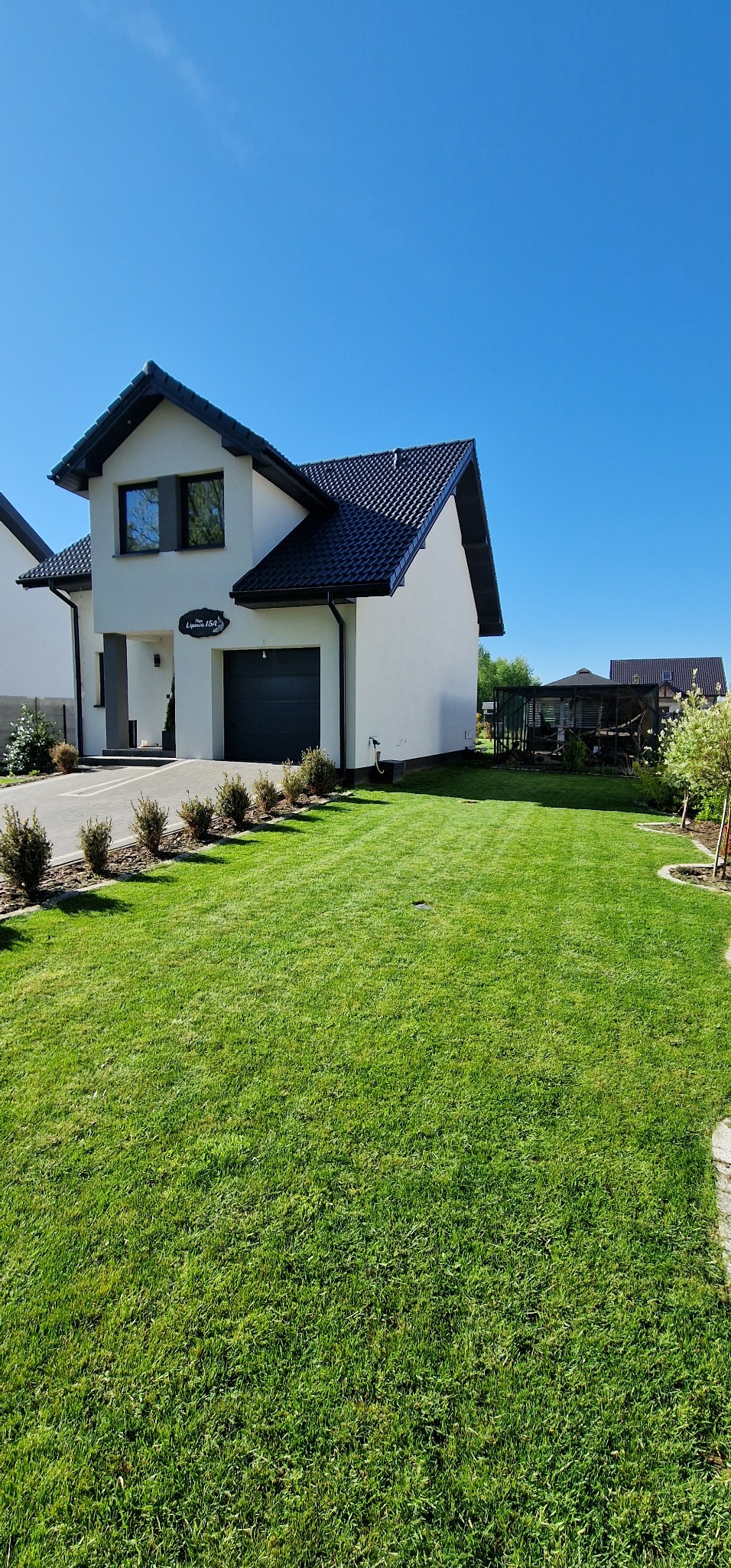 Zadbany trawnik przed nowoczesnym domem z ciemną stolarką okienną i garażem, w tle widoczna altana.