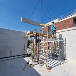 Pracownik na rusztowaniu kieruje dźwigiem podnoszącym betonowy element konstrukcyjny podczas budowy domu z białych bloczków, widoczne zbrojenia i elementy szalunku.