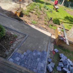 PHU Bartosz koper - Fragment układanej ścieżki z szarej kostki brukowej na przygotowanym podłożu, widoczny stos kostki obok, w tle trawnik, wózek z kostką i zaparkowany samochód dostawczy.