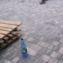 PHU Bartosz koper - Układanie szarej kostki brukowej na podwórku, widoczny fragment palety z kostką, butelka wody i pracownik ubijający kostkę