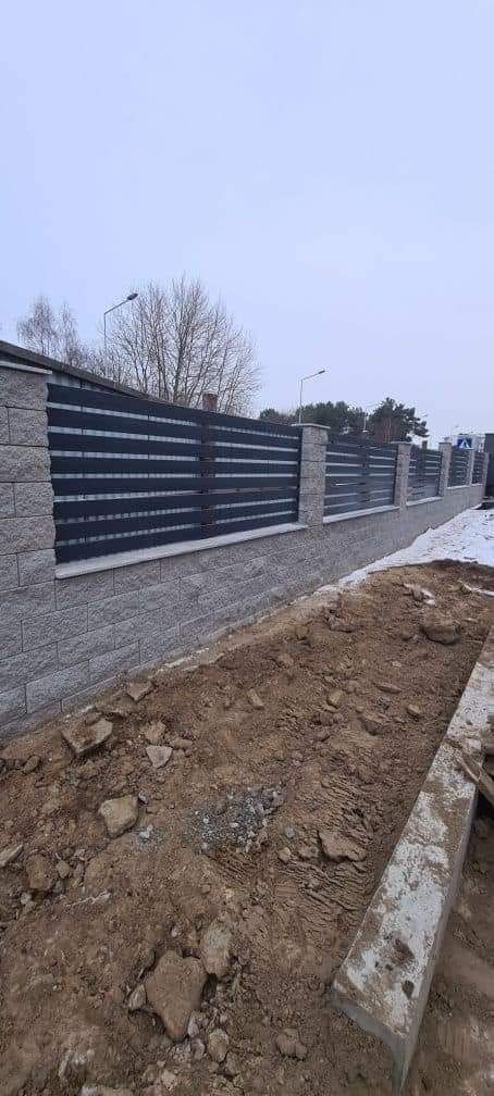 Nowoczesne ogrodzenie z szarej cegły z poziomymi, czarnymi panelami, widoczne świeżo przekopane podłoże i resztki śniegu.