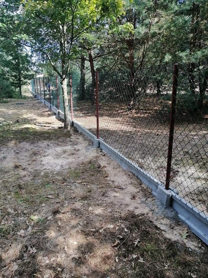 Długa, siatkowa ogrodzenie z betonową podmurówką i brązowymi słupkami na zalesionym terenie, widok z zewnątrz posesji.