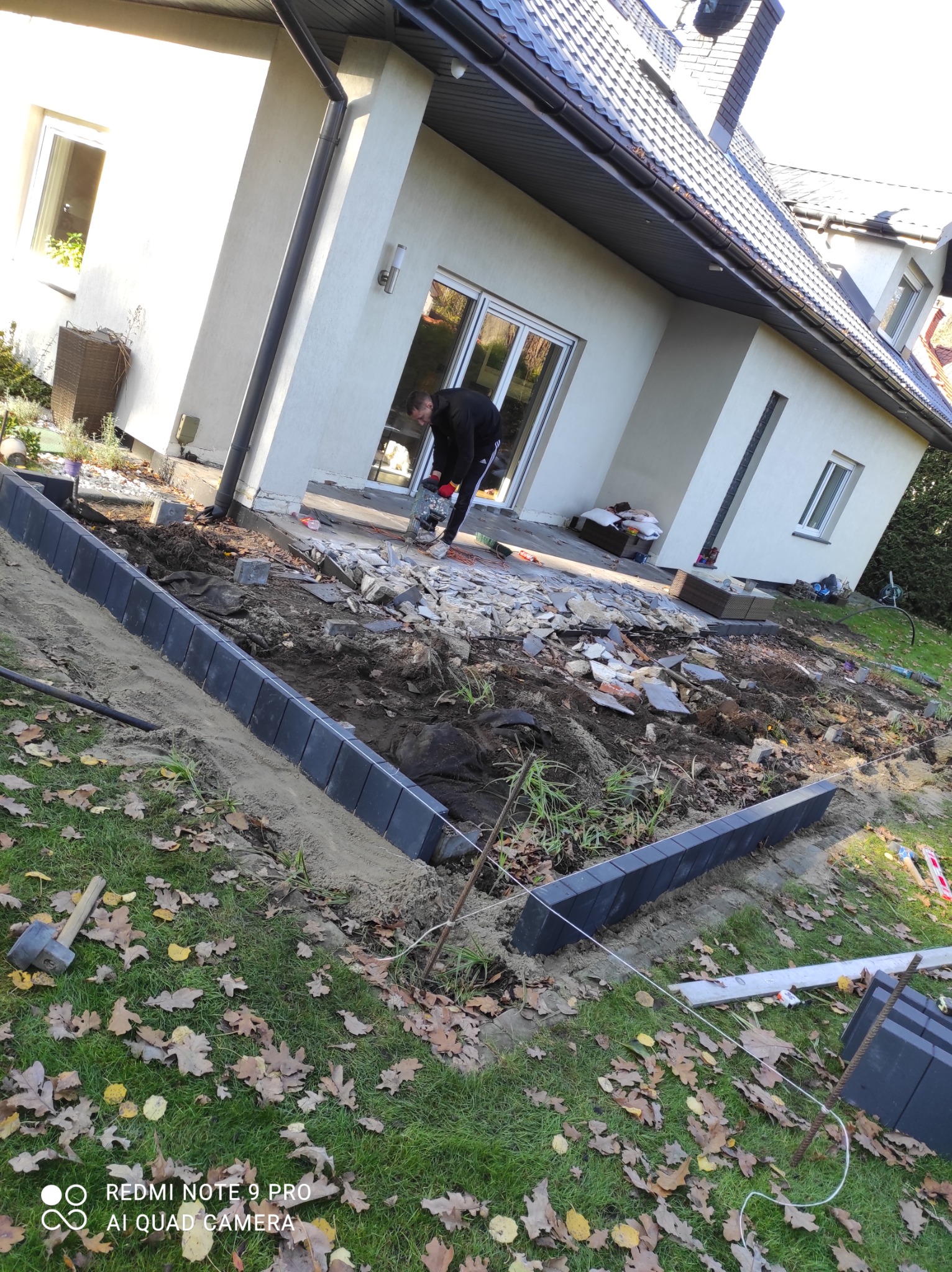 Układanie obrzeży z kostki brukowej na przygotowanym podłożu, widoczny pracownik używający przecinarki do kostki, w tle dom jednorodzinny.