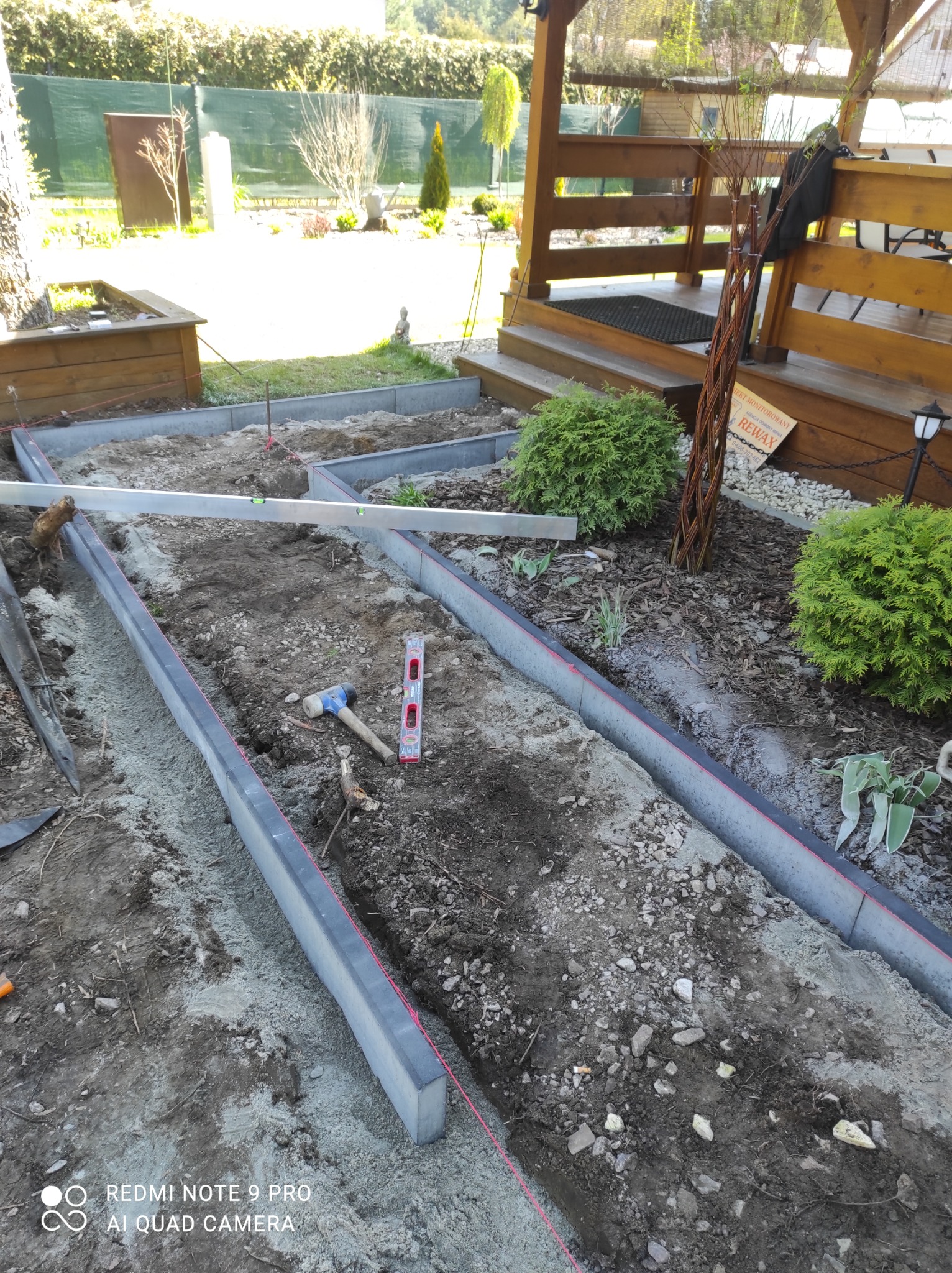 Przygotowanie podłoża pod ścieżkę z kostki brukowej: widoczne obrzeża, poziomica, gumowy młotek i sznurek wyznaczający linię. W tle drewniana altana i roślinność ogrodowa.