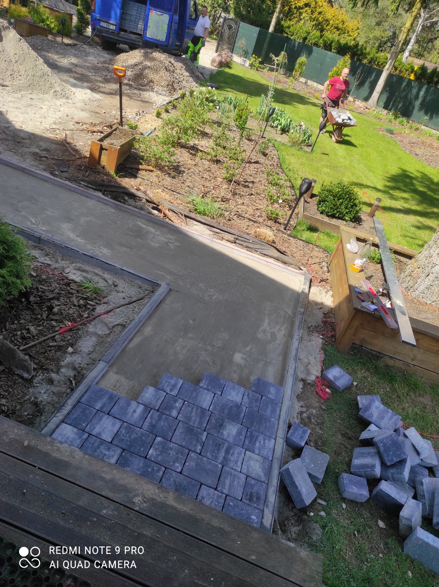 Fragment układanej ścieżki z szarej kostki brukowej na przygotowanym podłożu, widoczny stos kostki obok, w tle trawnik, wózek z kostką i zaparkowany samochód dostawczy.