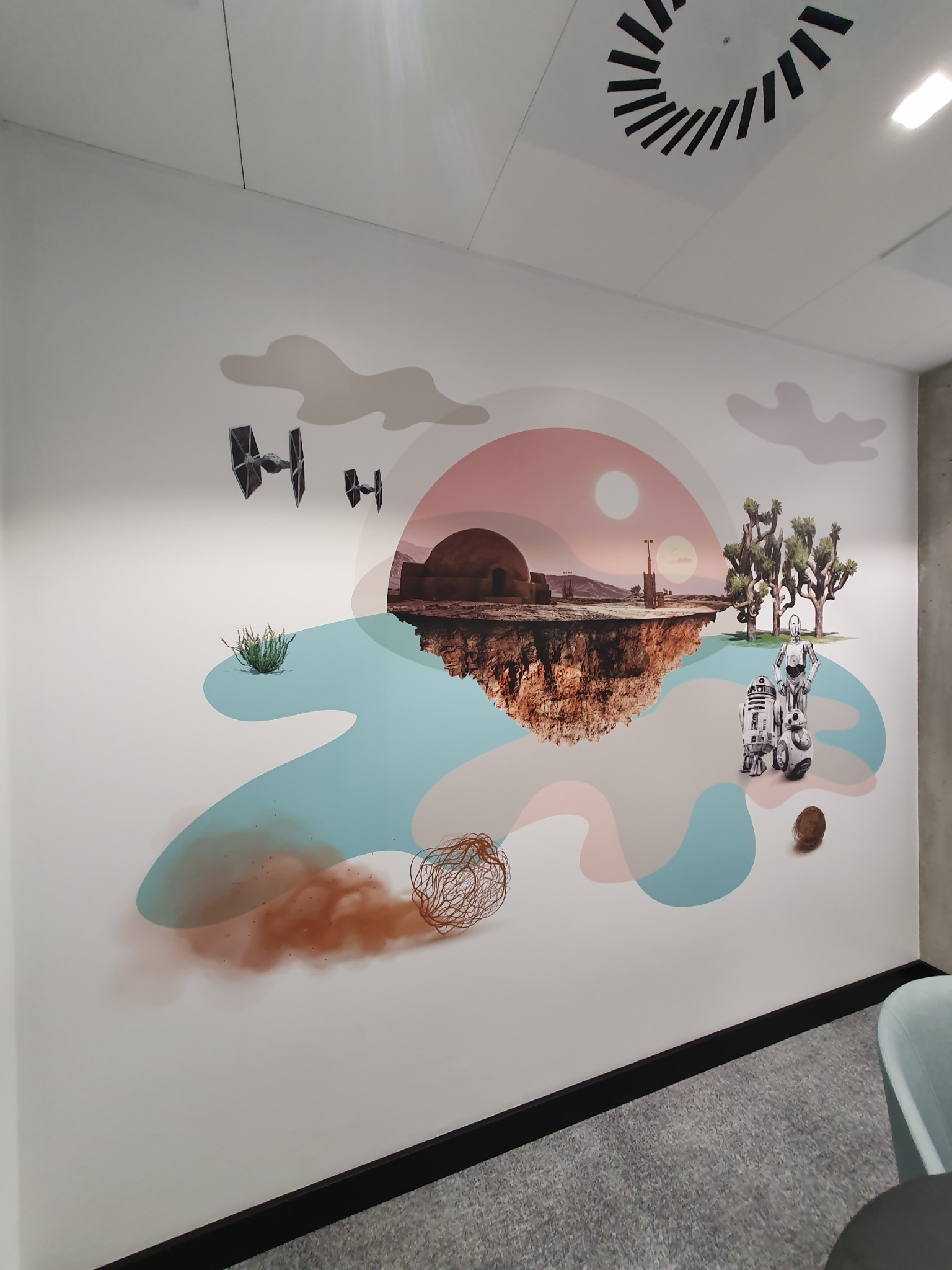 Ściana z fototapetą przedstawiającą pustynny krajobraz z elementami Star Wars: dom, statki kosmiczne, roboty, stylizowane chmury i plamy koloru.