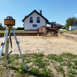 Wykonanie wykopu pod fundamenty domu jednorodzinnego, widoczny laser krzyżowy na statywie, mała koparka i pryzma ziemi.