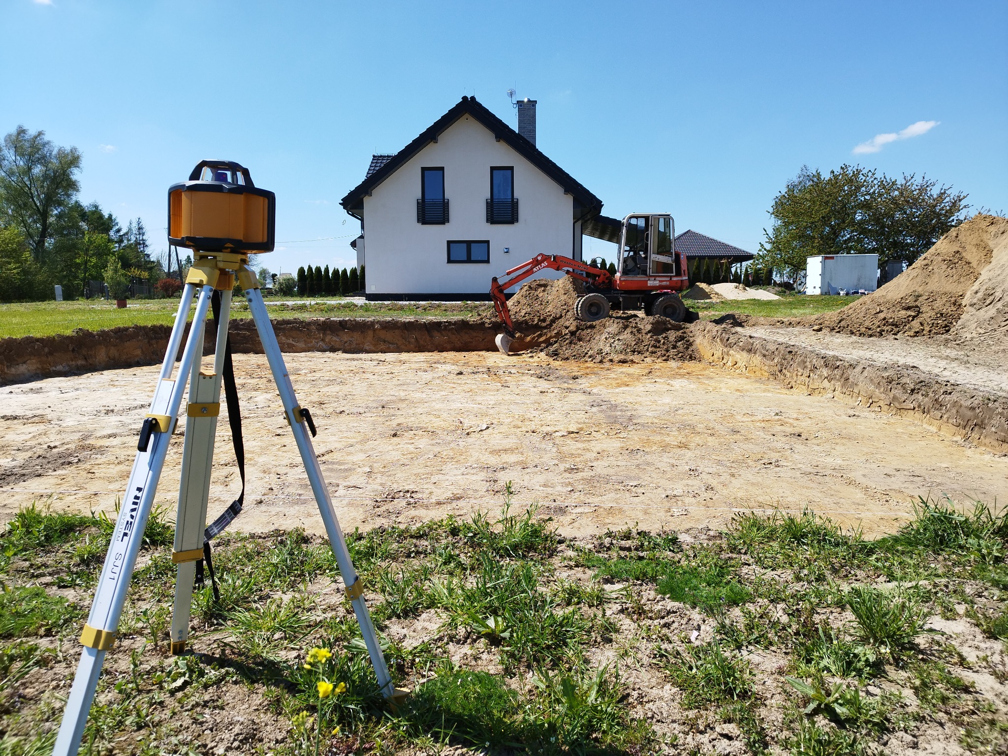 Wykonanie wykopu pod fundamenty domu jednorodzinnego, widoczny laser krzyżowy na statywie, mała koparka i pryzma ziemi.