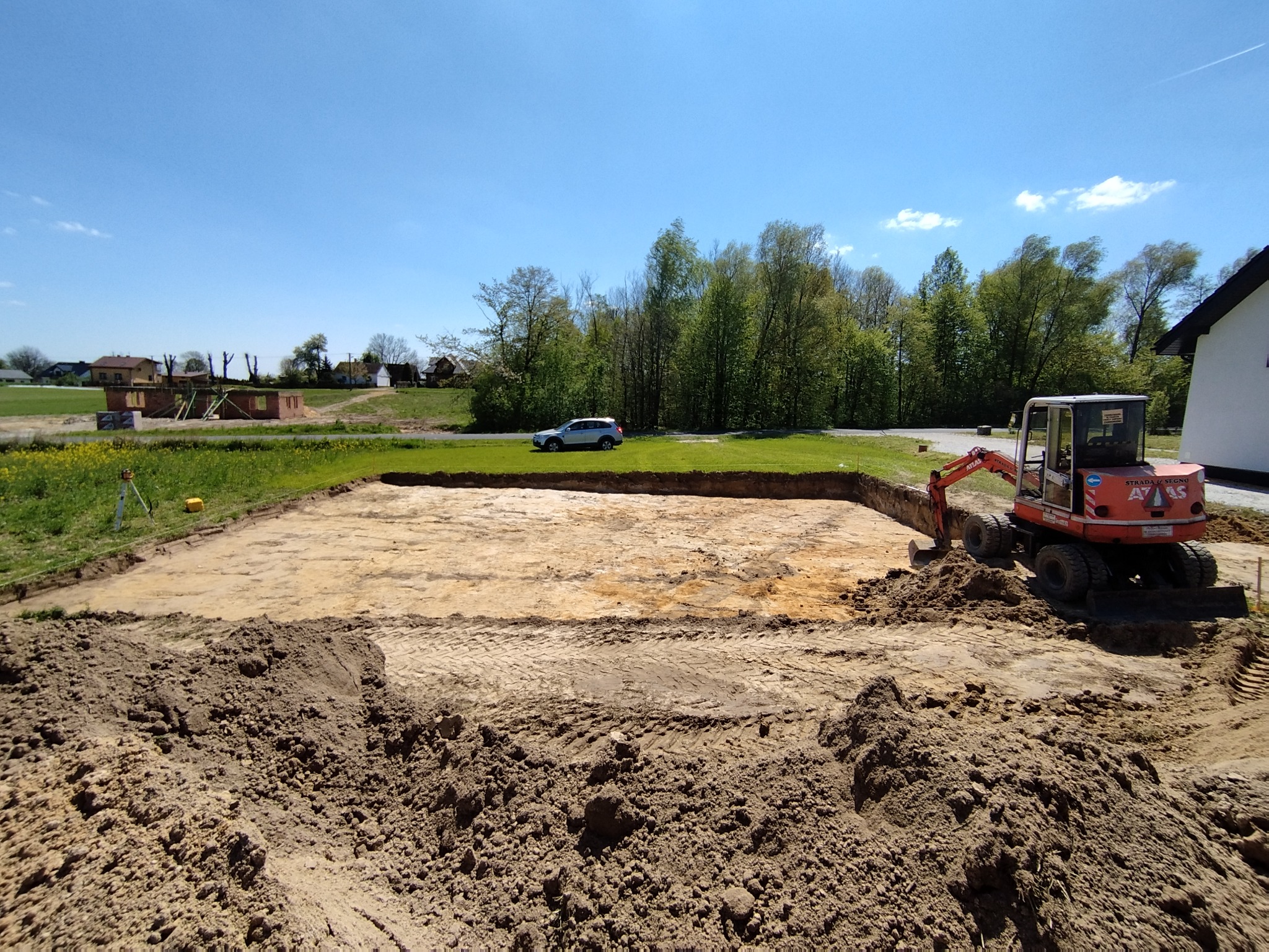 Wykopany fundament pod budynek, widoczny mały pomarańczowy koparka Atlas, wykopana ziemia oraz w oddali samochód osobowy na tle zieleni i błękitnego nieba.