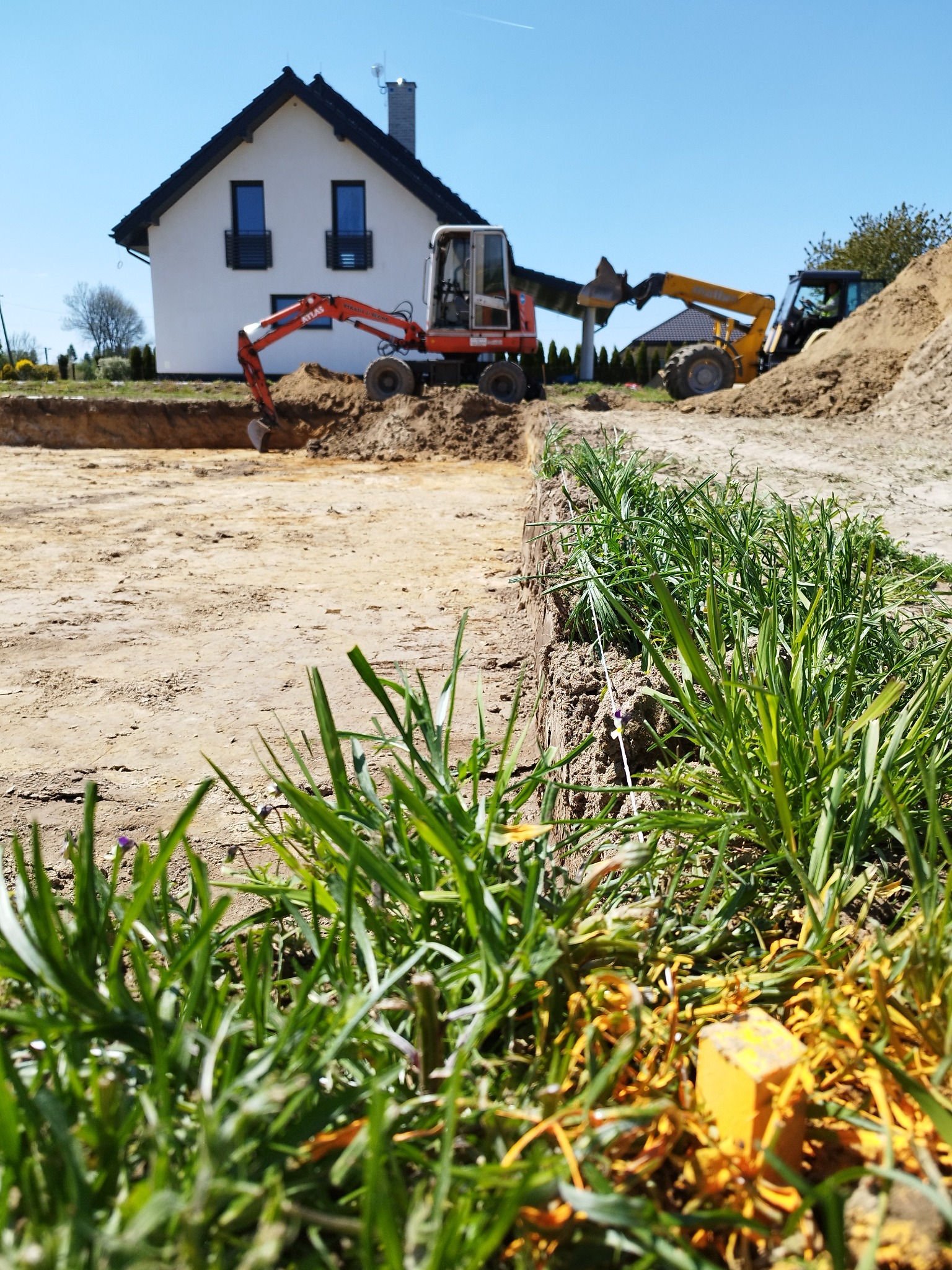 Wykop pod fundamenty domu jednorodzinnego z użyciem dwóch koparek, widoczne sznurki wyznaczające obrys, w tle biały dom z czarnym dachem.