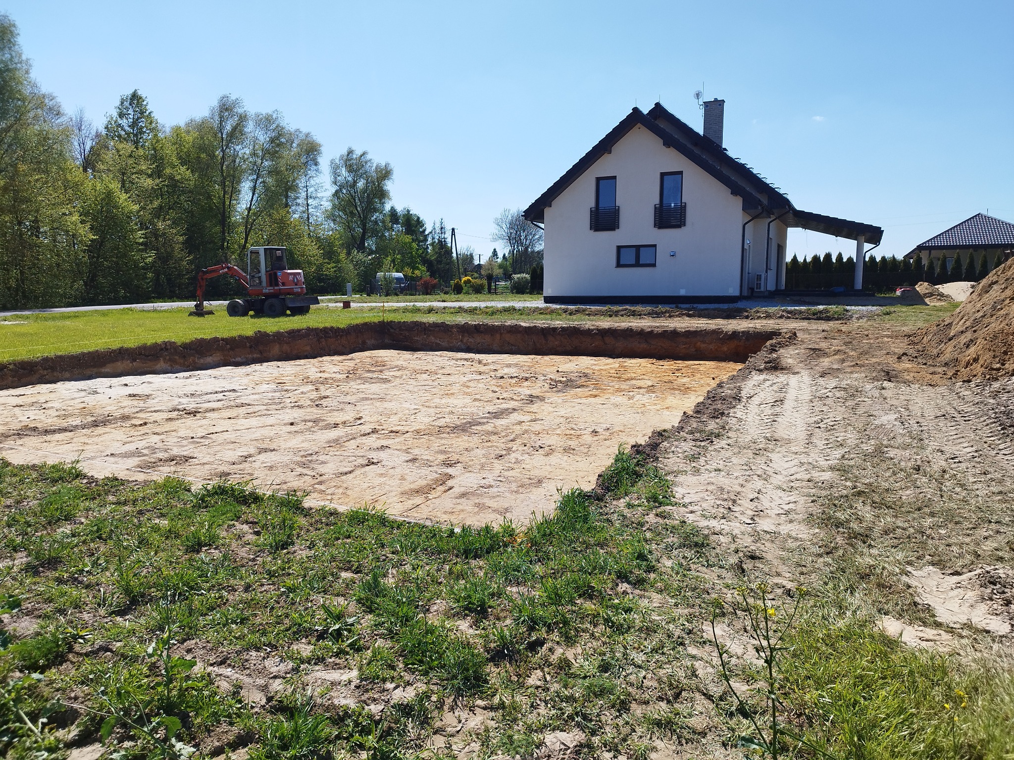 Przygotowany wykop pod fundamenty domu jednorodzinnego, widoczny mały czerwony traktor na trawniku, w tle dom z białymi ścianami i czarnym dachem, słoneczny dzień.