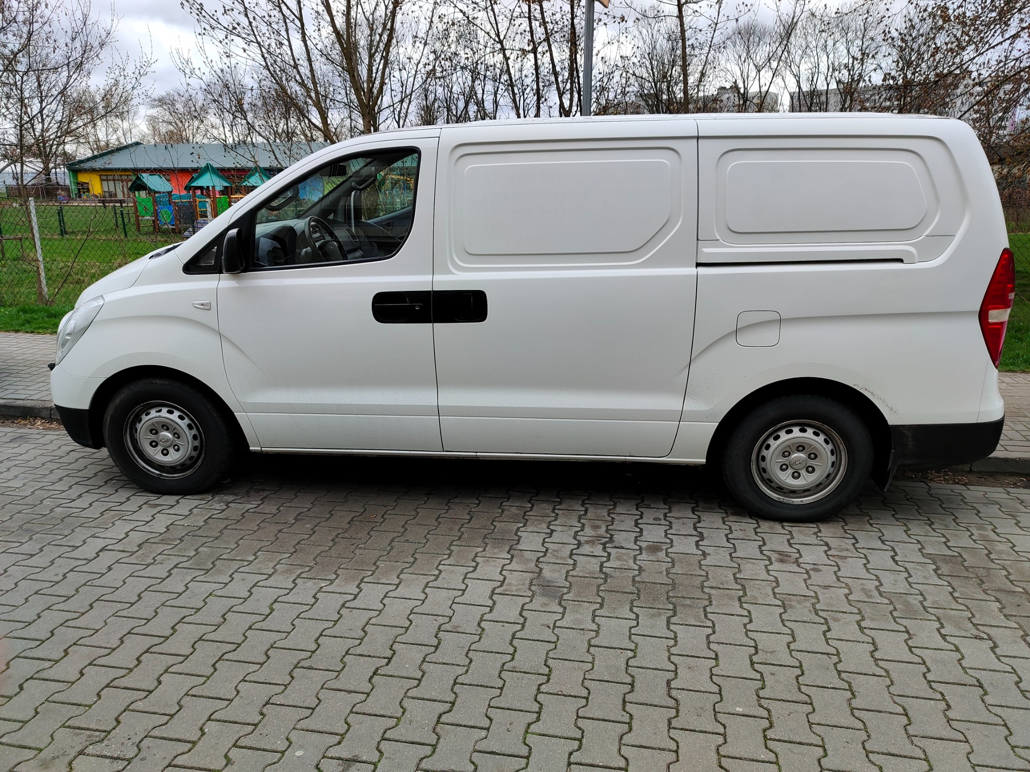 Biały furgon dostawczy zaparkowany na chodniku z kostki brukowej, w tle widoczny kolorowy budynek przedszkola i nagie drzewa.